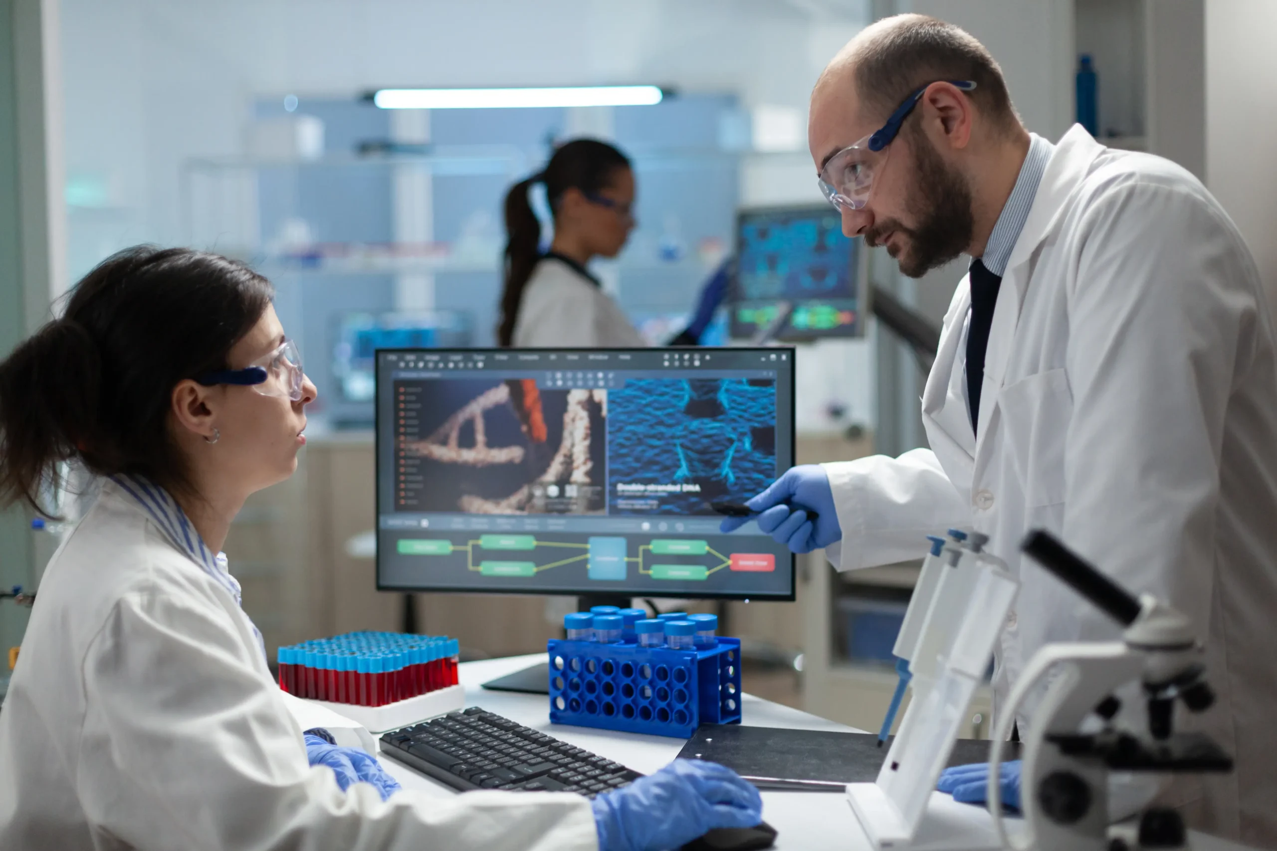 Researchers in a lab, wearing lab coats and safety goggles, discuss peptide-related DNA analysis in front of a computer screen.