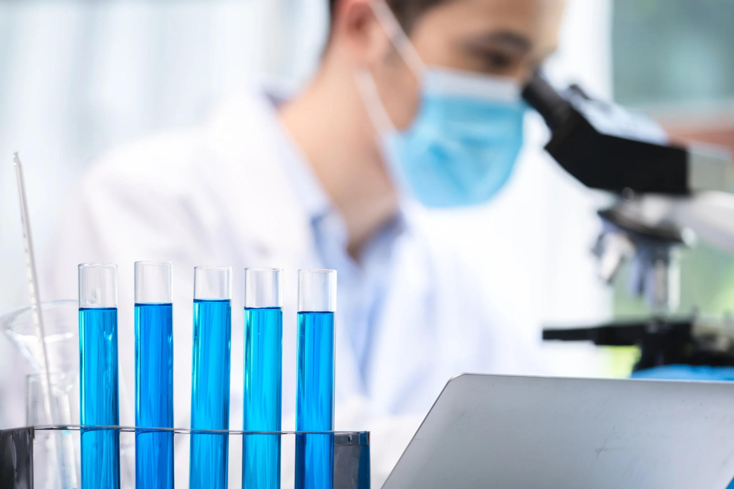 A set of test tubes filled with blue liquid in the foreground, with a scientist wearing a face mask using a microscope in the background. The scene represents peptide research in a laboratory.